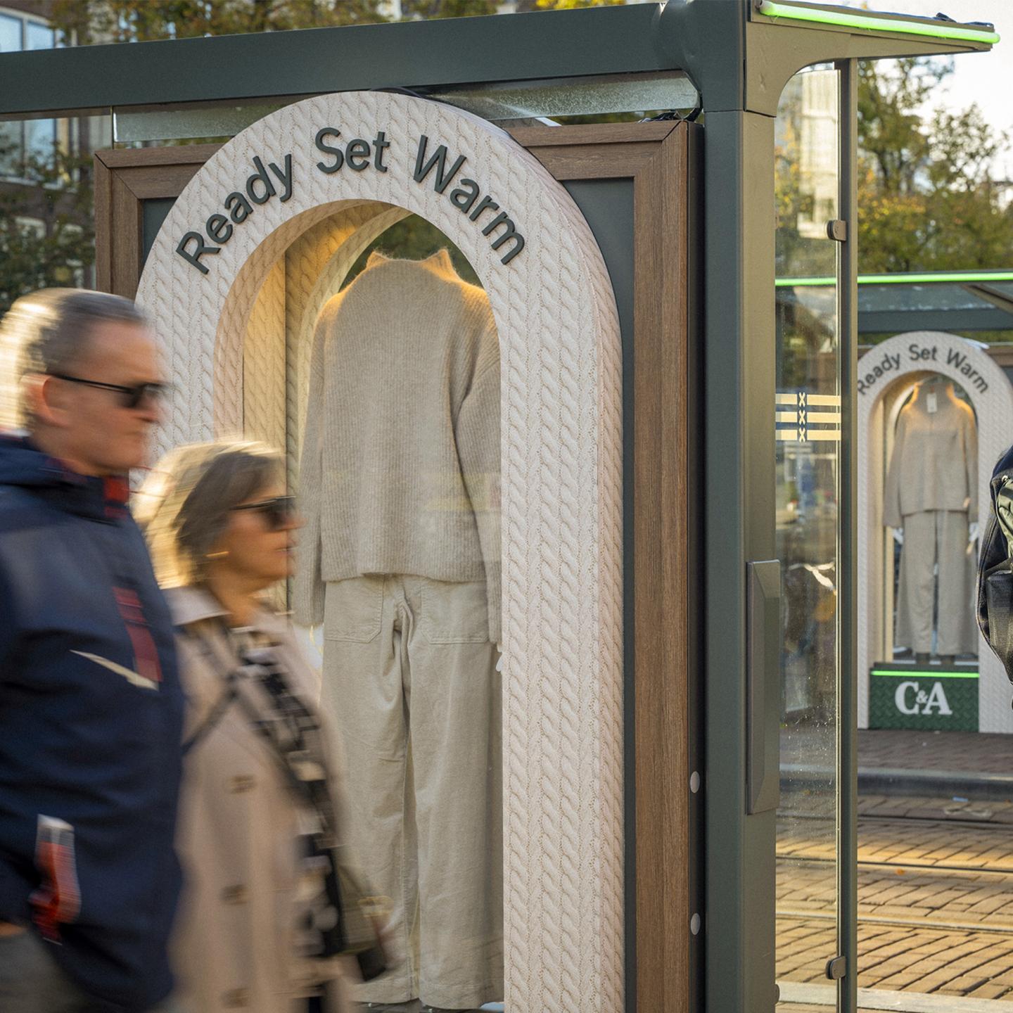 Close-up of a C&A knitwear display inside a wooden-framed Amsterdam bus shelter, with pedestrians passing in the foreground.