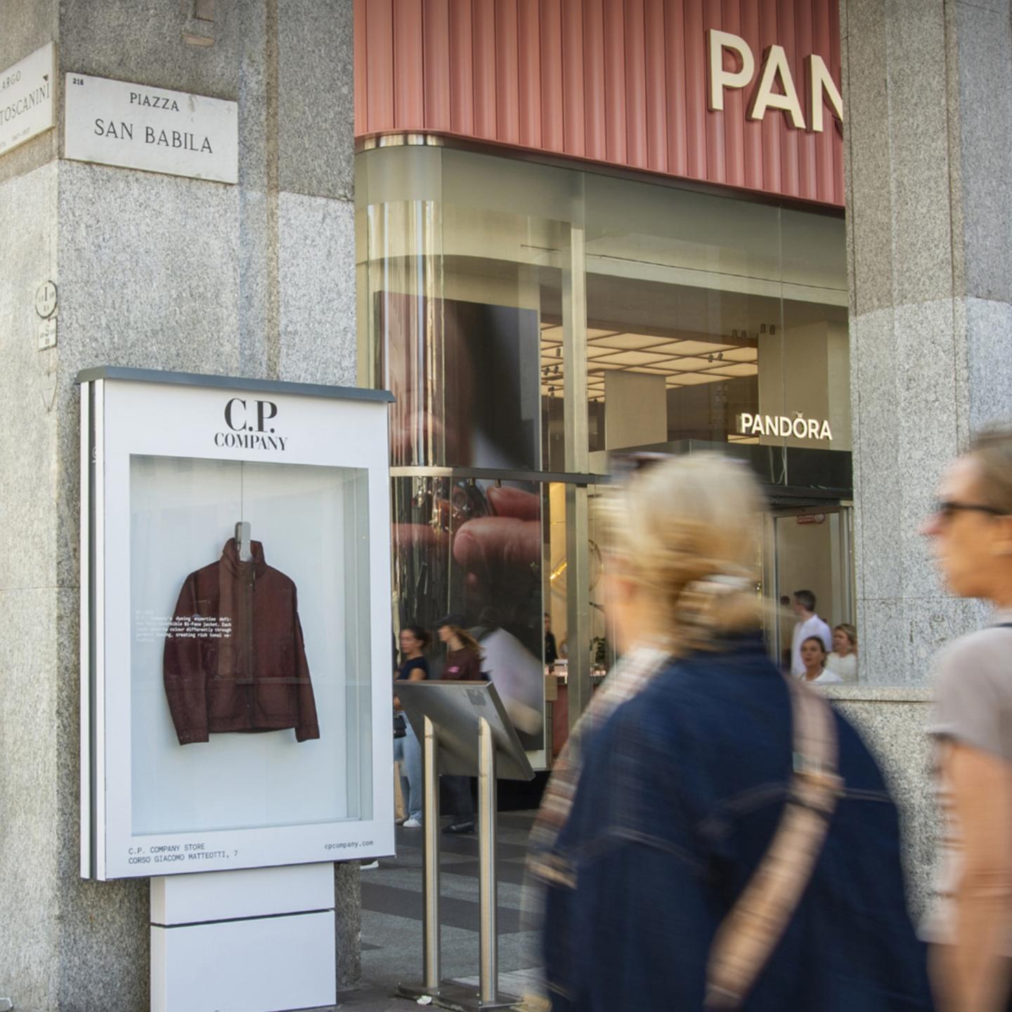 C.P. Company jacket displayed in an outdoor showcase at Piazza San Babila in Milan, with pedestrians passing by.