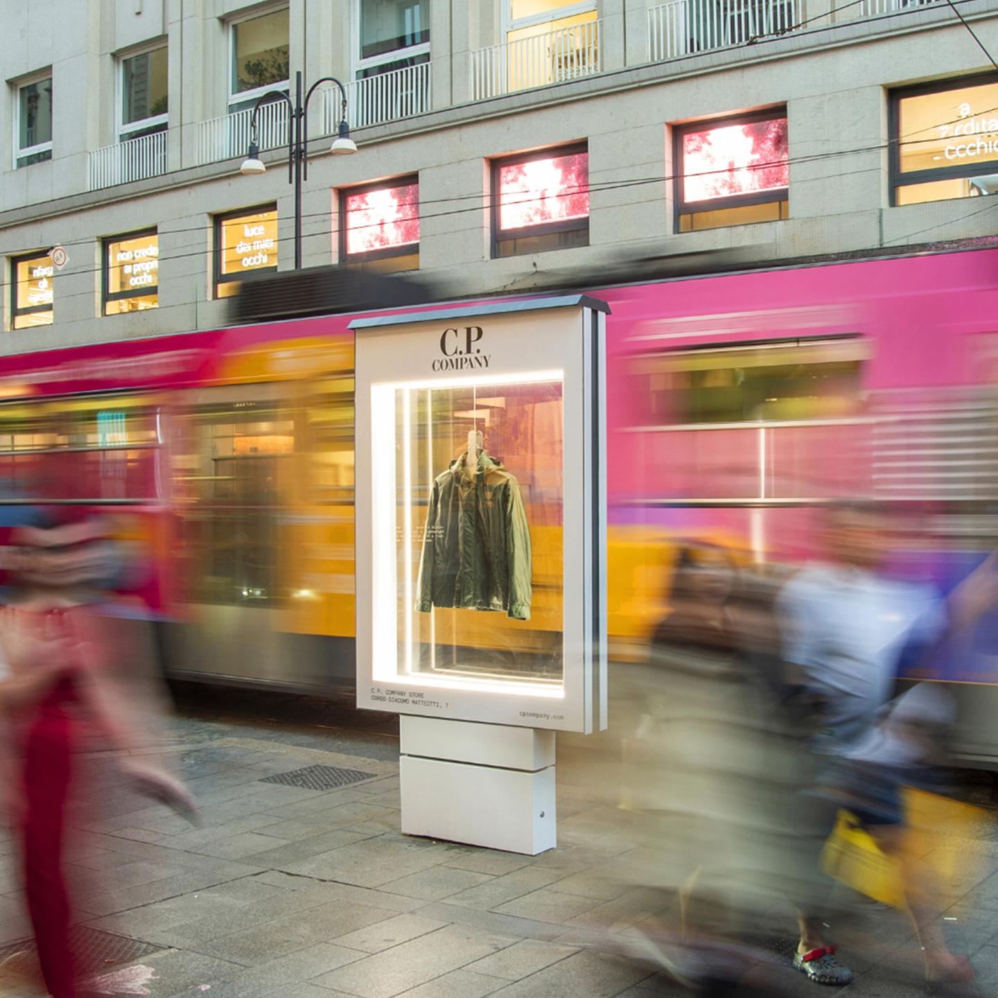 C.P. Company jacket displayed in an illuminated street showcase, with pedestrians walking past a tram stop in Milan.