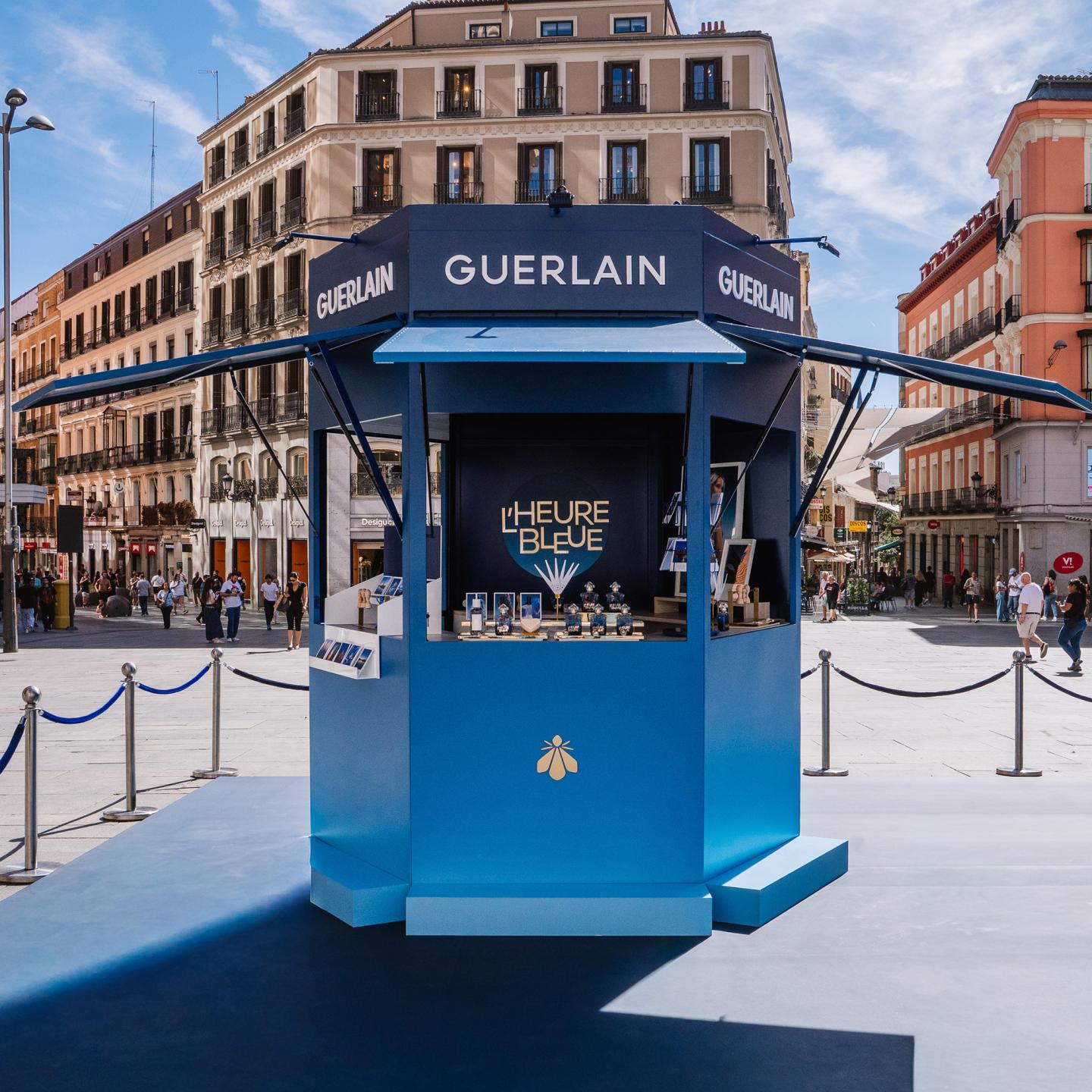 Front-facing view of the blue Guerlain L’Heure Bleue kiosk in the middle of Plaza Callao, surrounded by stanchions, with Madrid buildings in the background.