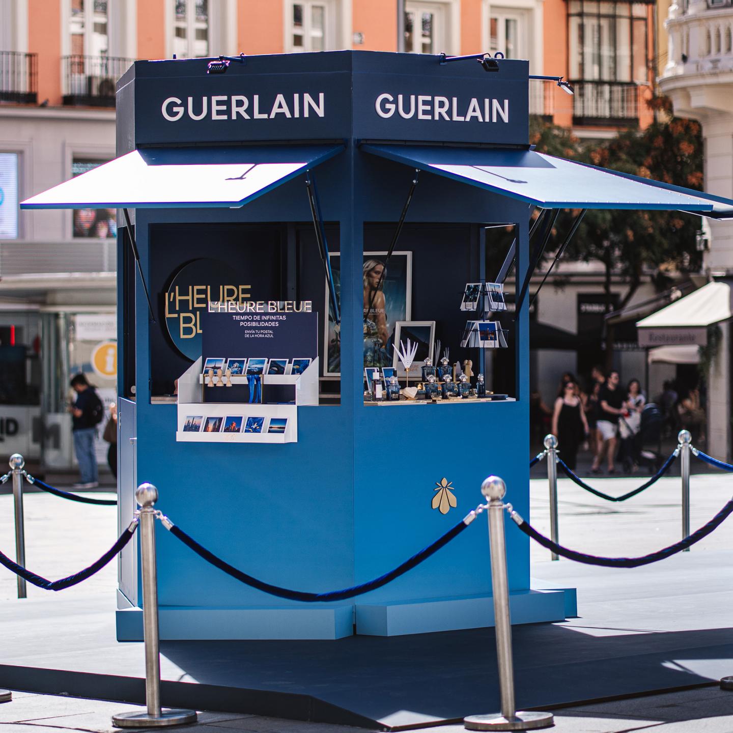 Side view of the blue Guerlain kiosk promoting L’Heure Bleue, with open awnings, product shelves and visitors in the background on Plaza Callao.