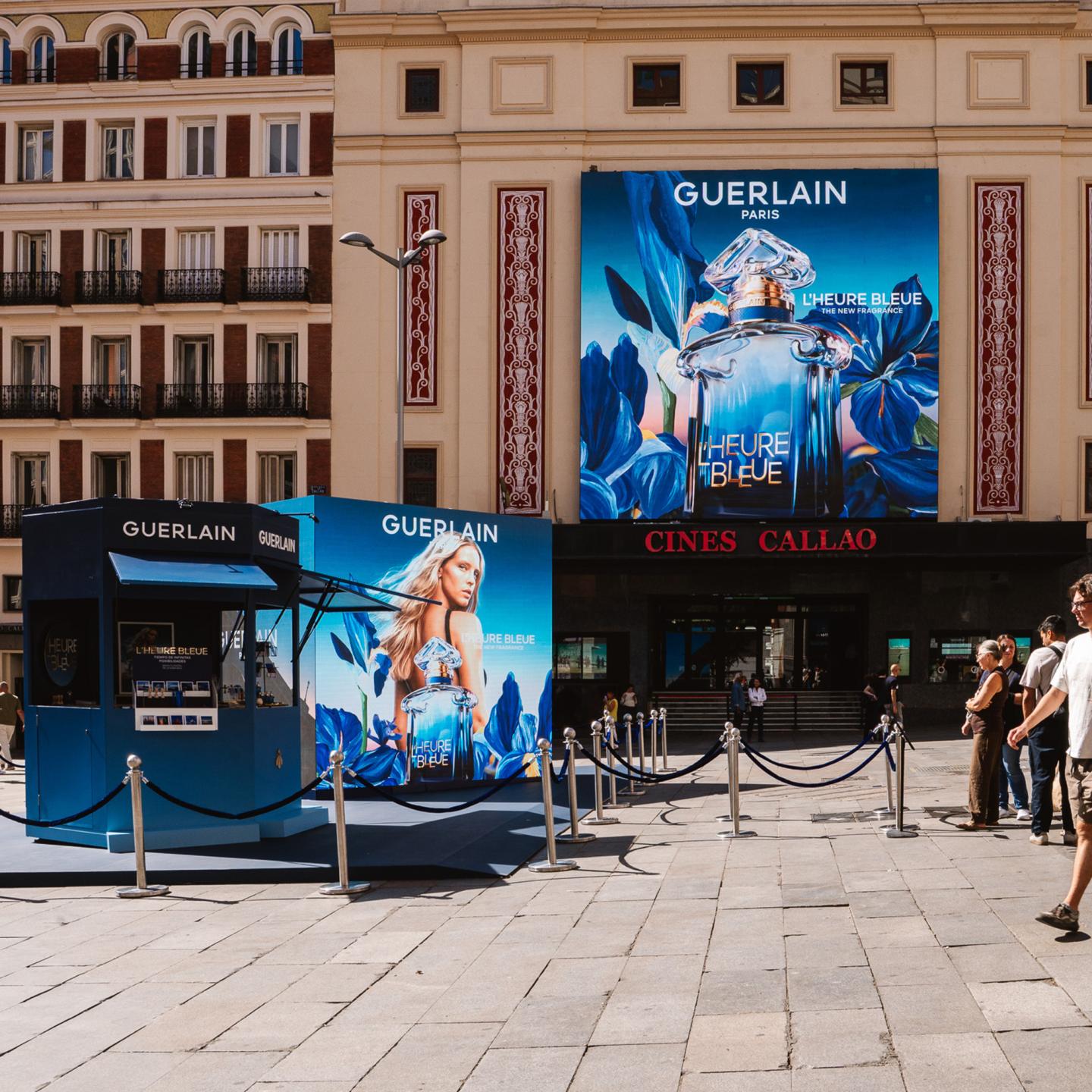Blue Guerlain L’Heure Bleue kiosk set up on Plaza Callao in Madrid, facing a large fragrance billboard on the cinema façade.