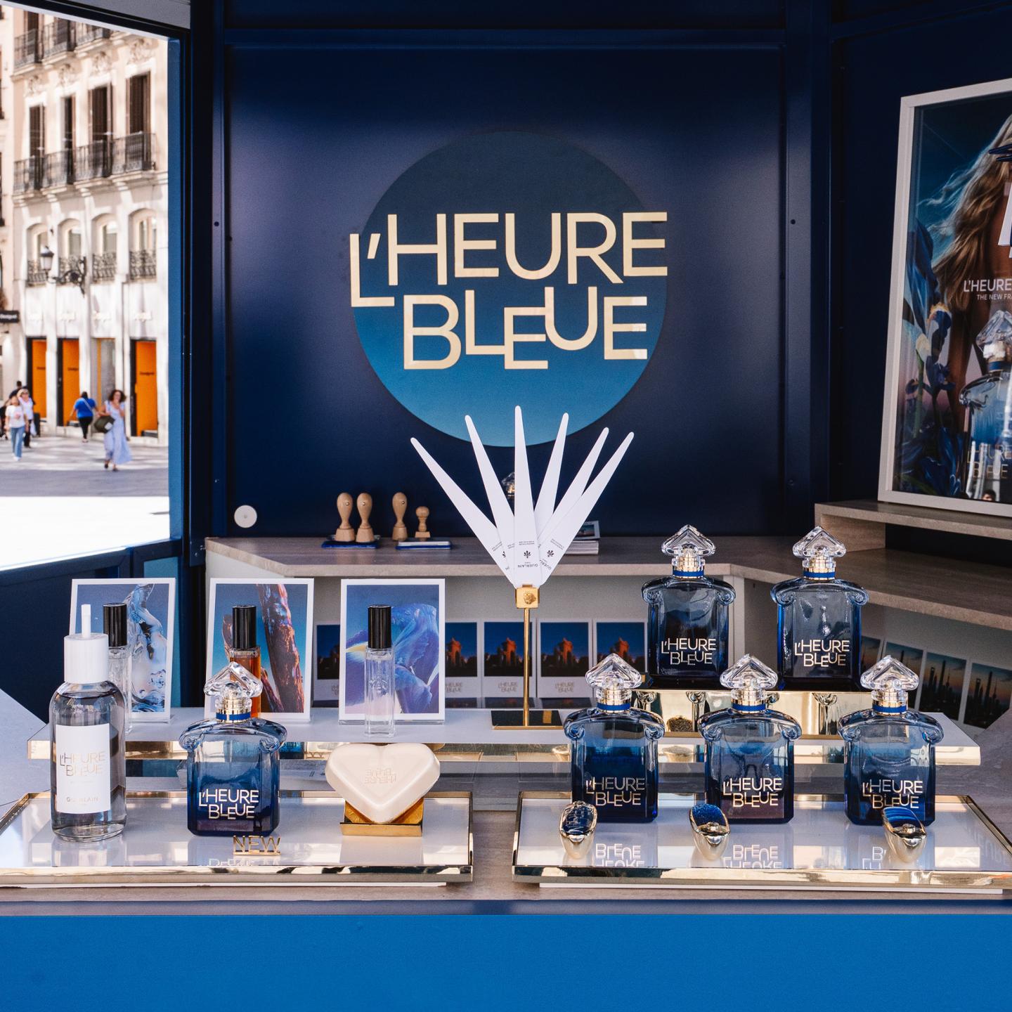 Inside the L’Heure Bleue kiosk, multiple Guerlain perfume bottles and fragrance testers are displayed against a dark blue backdrop.