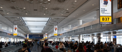 Passengers walk through Denver International Airport's concourse under synched digital screens displaying Alterra Mountain Co ads for Ikon Pass