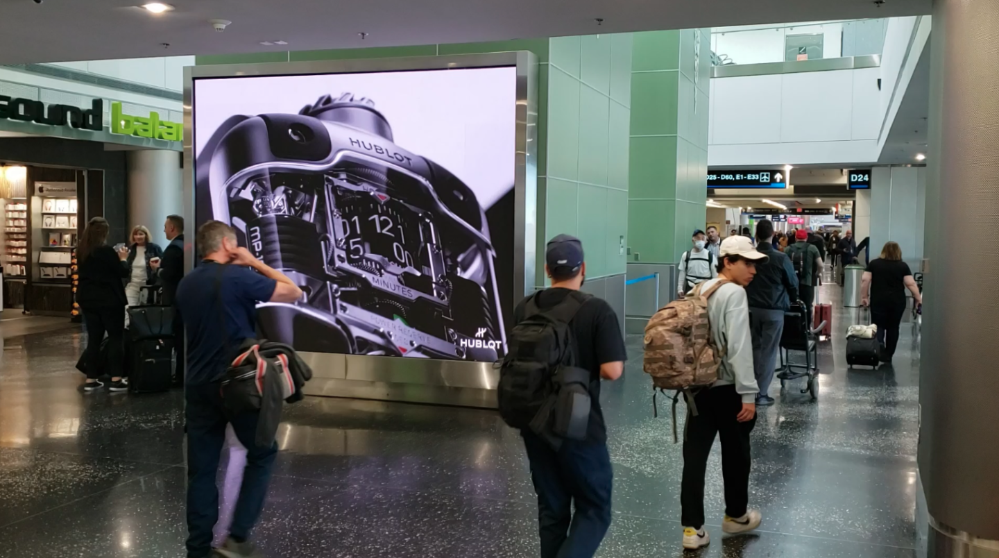 Passengers in airport concourse walk by large digital screen with Hublot watch advertising campaign