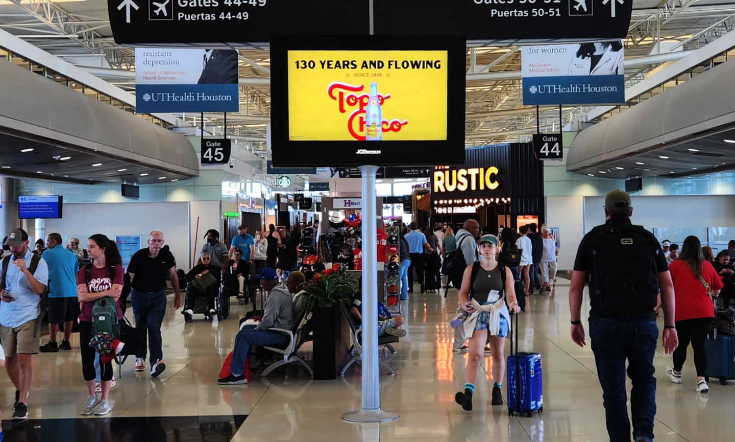 HOU concourse features a Topo Chico ad on a digital screen as passengers walk to and from gates on both sides