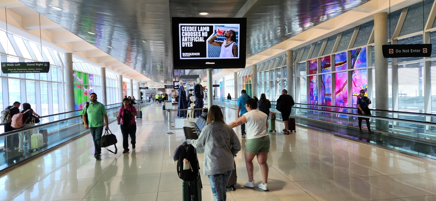 Passengers walk past prestige digital screen in Houston Hobby Airport displaying Ceedee Lamb on Coca Cola Body Armor ad
