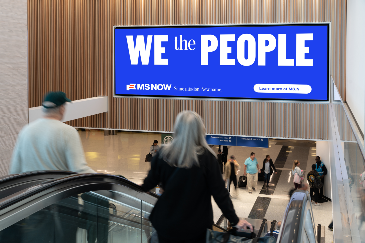 Arriving and departing passengers head down escalator in LAX Terminal 4 under large MS NOW digital OOH ad