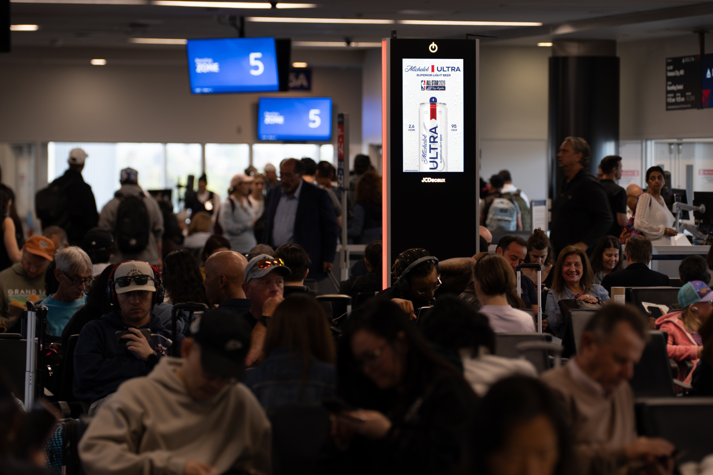 Travelers sit at gatehold area waiting for their flight. Charging station screen shows Michelob advertising for the NBA All Star Game