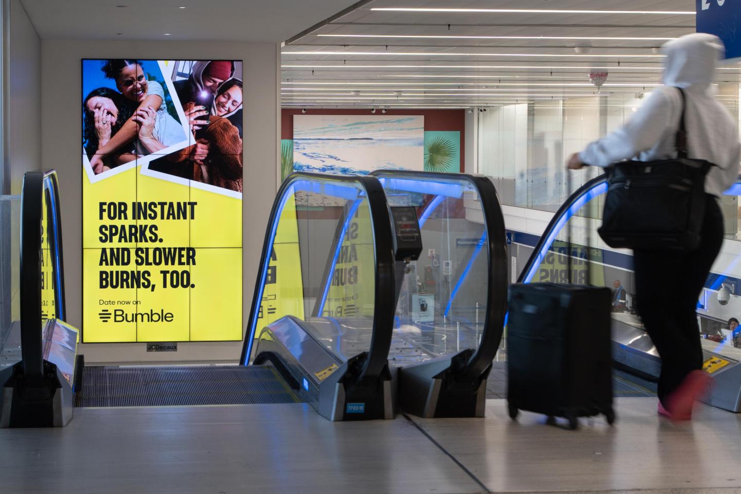 Passengers head down an escalator past a large digital advertisement for Bumble in LAX