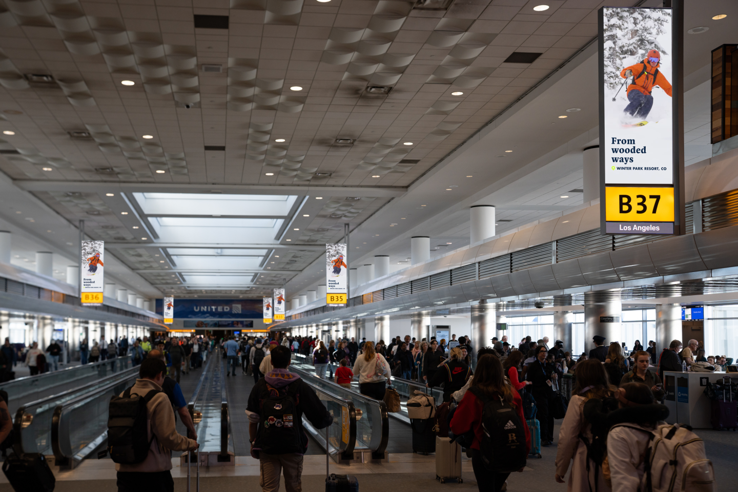 Passengers walk through Denver International Airport's concourse under synched digital screens displaying Alterra Mountain Co ads for Ikon Pass