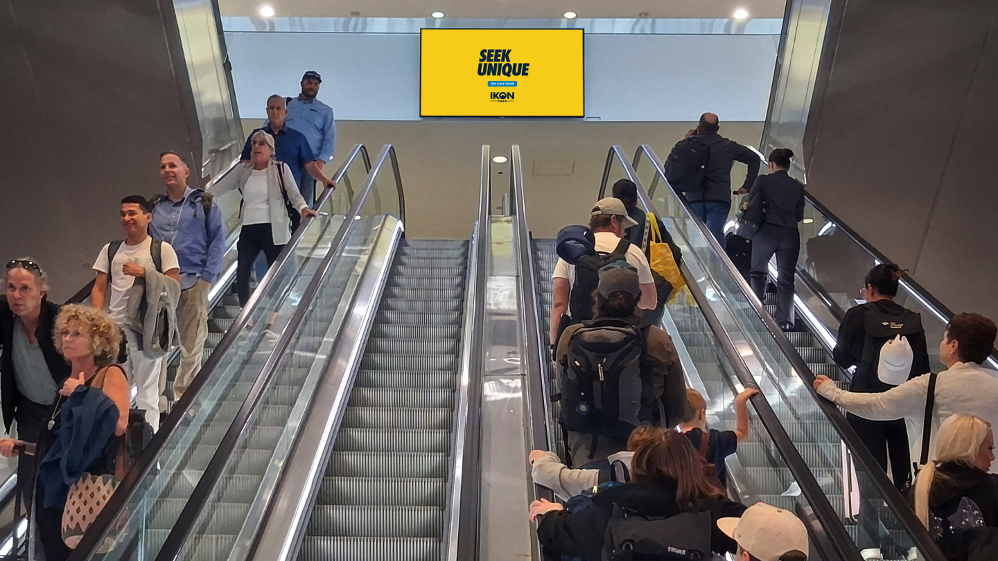 Passengers head up and down escalator passing a large format digital spectacular displaying Ikon Pass advertising in DEN